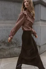 Blusa de manga larga con estampado de cuadros vichy en tonos burdeos y beige. Presenta cuello mao y grandes volantes fruncidos que cruzan el pecho y los hombros. Los puños son elásticos y fruncidos.