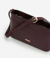 Cartera bandolera pequeña de formato rectangular, confeccionada en poliuretano con acabado liso. Presenta una correa larga ajustable, cierre superior con cremallera y un pequeño aplique metálico dorado con el logo de la marca en el frente.