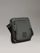 Bolso tipo reportero o bandolera de Calvin Klein, color verde oliva oscuro con detalles en marrón oscuro. Presenta un diseño funcional con un bolsillo exterior frontal con cierre y un parche de cuero sintético con el logo de la marca. Incluye correa ajustable de tela.