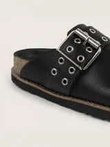 Zueco de cuero negro con diseño de punta redonda y tira ajustable en el empeine con hebilla metálica y ojales decorativos. Interior forrado en cuero y suela con vira reforzada.
