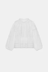 Blusa de cuello subido y manga larga con puños elásticos. Presenta un diseño con bordados florales y detalles de puntilla a tono, con un corte que incluye volantes en el bajo.