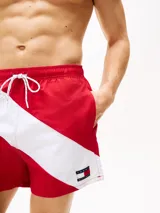 Short de baño rojo con cintura elástica ajustable con cordón, corte amplio y logo de Tommy Hilfiger en la pierna. Presenta una franja diagonal blanca con el logo de la marca en la parte inferior.