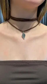 Gargantilla estilo choker de doble vuelta confeccionada en cordón de gamuza marrón, con un dije metálico plateado en forma de hoja.