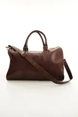 Bolso de viaje color marrón, confeccionado en cuero vacuno, con doble asa de mano y correa ajustable y desmontable. Cuenta con herrajes metálicos y costuras reforzadas.