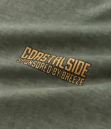 Remera de hombre, modelo comfort, color verde oliva desteñido, con cuello redondo y manga corta. Presenta un pequeño estampado en el pecho con lettering en tonos naranjas que dice "COASTALSIDE".