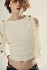 Jersey de punto blanco marfil, con cuello barco y mangas anchas tipo capa decoradas con botones a lo largo de los hombros y brazos.