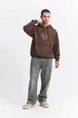 Canguro oversize color marrón, con capucha, cordones ajustables, bolsillo frontal con cierre y estampa frontal con texto y logo de la marca.