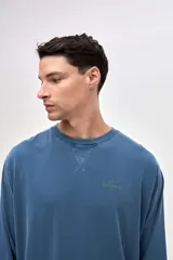 Remera de manga larga color beige, confeccionada en algodón lavado con textura suave. Presenta cuello redondo con detalle de costura en V, corte clásico y estampado pequeño en el pecho con la palabra Uniform.