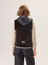 Chaleco gris con cuello alto y capucha, cierre frontal con botones a presión. La parte delantera es de lana gris lisa y la parte interior del cuello y el forro son de tela con patrón de espiga en blanco y negro.