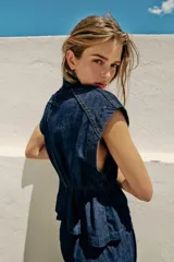 Chaleco de denim azul oscuro con cuello alto, manga sisa y cintura ajustable con elástico. Cierre frontal con botones a presión metálicos.