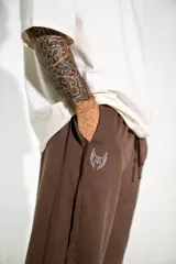 Pantalón de estilo urbano color marrón, con cintura elástica ajustable mediante cordón, bolsillos laterales y bajos ajustables con tiras. Presenta un pequeño estampado en la pierna.