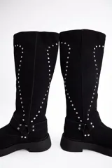 Bota de caña alta confeccionada en cuero vacuno, color negro, con diseño de tachas metálicas dispuestas en forma de arco a lo largo de la caña y correa decorativa con hebilla en la zona del tobillo. Presenta suela track de goma robusta.