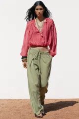 Pantalón ancho de tiro alto color beige claro, con cintura elástica, cordón ajustable y pinzas delanteras. Presenta bolsillos frontales y bolsillos de plastrón en la espalda. Incluye un cinturón del mismo tejido con apliques metálicos.