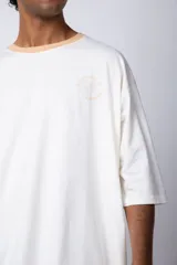 Remera de corte oversize en color beige, con cuello redondo en contraste negro y una manga en color negro. Presenta un pequeño estampado circular en el pecho con la frase 'Don't worry, be happy'.