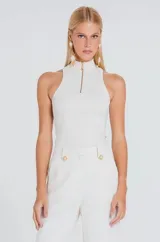 Musculosa de tejido canelado en color blanco, con cuello alto y cierre frontal parcial con cremallera dorada. Presenta un diseño ajustado al cuerpo y sisas amplias.