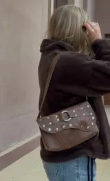 Cartera de hombro color marrón con acabado texturizado, solapa frontal decorada con tachas metálicas plateadas y una argolla central. Cuenta con correa ajustable y cierre principal.