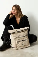 Mochila shopper color beige, confeccionada en poliéster. Presenta un diseño versátil con múltiples cierres, compartimento inferior expandible y un bolsillo frontal removible que funciona como estuche. Incluye correas ajustables y removibles para usar como mochila o bandolera, además de asas fijas para llevar al hombro.