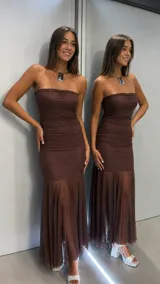 Vestido largo color chocolate, con diseño strapless y cuerpo ajustado con frunces laterales. La falda presenta un corte sirena con un panel inferior de tela transparente y fluida.