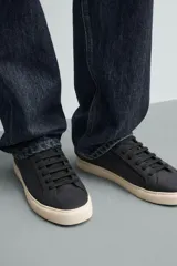 Zapatilla de estilo minimalista en color negro, con cordones al tono, suela de goma en color crema y detalle de talón en contraste.