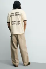 Camiseta de corte regular, color beige con cuello y bordes de mangas en color marrón oscuro. Presenta un diseño de texto bordado en el pecho con la frase 'Letting Time Pass By'.