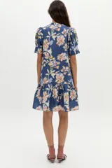 Vestido corto azul de lino con estampado floral en tonos rosados, beige y celestes, cuello mao con escote en V, mangas cortas abullonadas, botones frontales ocultos y volado en el ruedo.