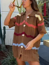 Camisa de tejido de punto con diseño oversize, cuello tipo polo y cierre frontal con botones. Presenta un patrón de rayas horizontales en zigzag de colores contrastantes sobre una base marrón.