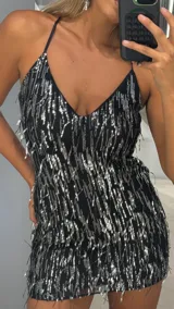 Vestido corto de fiesta con escote en V y tirantes finos. Presenta una base negra cubierta con flecos de lentejuelas plateadas que crean un efecto brillante y dinámico.