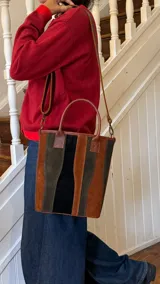 Bolso tipo matera confeccionado en cuero, con diseño de paneles verticales en tonos marrón, verde oliva y negro. Cuenta con doble asa de mano y correa larga ajustable y extraíble para llevar al hombro.