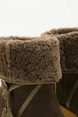 Bota de caña media color verde oliva, confeccionada en cuero con detalle de solapa superior en lana rizada. Presenta un cordón decorativo con borlas en el mismo tono y una suela de plataforma tipo cuña en color beige.