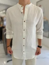 Camisa de lino color blanco, de manga larga con puños vueltos y cuello mao. Cuenta con cierre frontal de botones marrones.