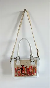 Cartera Diri de cuero natural uruguayo, en tonos marfil y arena, con intervención artística pintada a mano en el frente con pinceladas en rojos, terracotas y dorados. Cuenta con estructura firme, herrajes metálicos en acabado oro viejo, cierres, manijas ergonómicas y correa regulable desmontable.