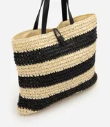 Cartera tipo saco shopper confeccionada en paja trenzada, con diseño de rayas en color natural y negro. Cuenta con asas de hombro de material sintético y cierre con cordón ajustable con detalle de concha.