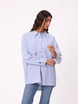 Camisa de corte oversize con diseño a rayas verticales en tonos azul y blanco. Presenta cuello clásico, cierre frontal con botones y un bolsillo en el pecho con detalle de etiqueta.