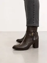 Bota de caña media confeccionada en cuero color marrón oscuro, con punta almendrada y taco cuadrado de 7 cm. Presenta una tira decorativa con hebilla metálica dorada en la zona del tobillo.