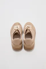 Zapatillas urbanas de corte bajo en tonos beige y tostado, combinando paneles de cuerina y gamuza sintética. Cuentan con cordones anchos tipo cinta de raso en color crudo, pespuntes decorativos y suela de goma al tono.