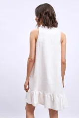 Vestido corto blanco sin mangas, con escote en V y detalle de broche dorado en el centro. La falda tiene un volado en el ruedo.