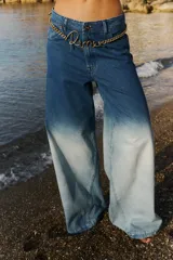 Jean de tiro medio con corte oversize y botamanga ancha. Presenta un diseño de lavado degradado que va de un azul intenso en la cintura a un tono claro en la parte inferior, con efecto gastado. Incluye avíos metálicos y badana de PU.