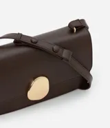 Cartera tipo shoulder bag de tamaño mediano, confeccionada en poliuretano color marrón oscuro. Presenta una solapa frontal con un detalle metálico dorado de forma orgánica y asa ajustable.