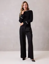 Buzo de tejido soft color negro, con cuello a la base y mangas largas. Presenta apliques de perlas decorativas en el contorno del cuello y en los puños.