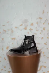 Botineta de cuero charol negro con acabado brillante, diseño de punta fina y taco cuadrado. Presenta un panel elástico lateral y una correa decorativa con hebilla metálica plateada.