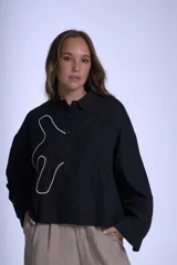 Camisa negra de manga larga con cuello clásico y cierre frontal con botones. Presenta bordados abstractos en blanco y negro en el frente.
