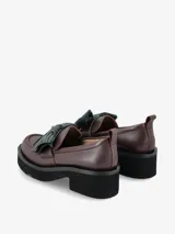 Mocasín de cuero negro con plataforma gruesa y taco cuadrado. Presenta un detalle decorativo de lazo plisado en color azul oscuro sobre el empeine y una pequeña tira en el talón.