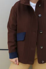 Campera de paño de lana en color beige jaspeado, con diseño oversize. Presenta detalles en pana color marrón chocolate en los puños y en las tapas de los bolsillos frontales. Cuenta con cierre frontal de botones grandes y cuello alto.