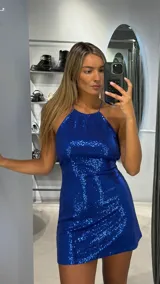 Vestido corto de fiesta confeccionado en tela con lentejuelas azules. Presenta un diseño ajustado al cuerpo con escote halter y espalda descubierta.