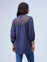 Blusa color beige de gasa con cuello a la base, escote en V y abertura central con botones. Presenta detalles de crochet en hombros y espalda, mangas 3/4 amplias con puño y botón. El largo de la espalda es mayor al delantero.