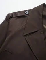 Chaqueta corta de color marrón chocolate, con diseño de doble botonadura, solapas amplias y charreteras en los hombros. Presenta mangas largas con puños ajustables mediante trabillas y bolsillos laterales integrados.