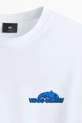 Remera blanca de punto de algodón, cuello redondo, mangas cortas y hombros caídos con pequeño estampado azul en el pecho.