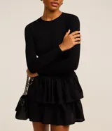 Vestido corto negro de viscosa con mangas largas, cuello redondo y falda godê con superposición de volantes. La parte superior es acanalada y la inferior tiene capas fruncidas.