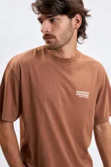Remera de manga corta color marrón con efecto lavado, cuello redondo y estampa pequeña en el pecho con diseño de tablero de ajedrez y texto.
