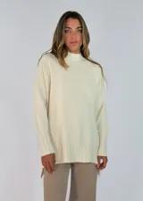 Sweater de punto color crema, con cuello alto, mangas largas y puños con detalle de tejido trenzado. Presenta un diseño holgado con ruedo asimétrico y aberturas laterales.